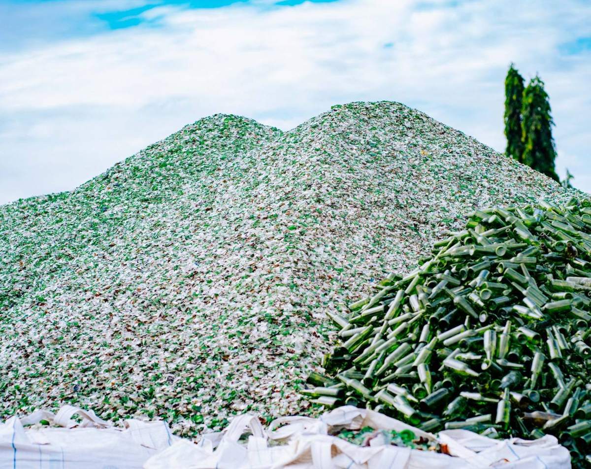 Movilă de cioburi de sticlă verde și albă, cu o grămadă de sticle întregi alături