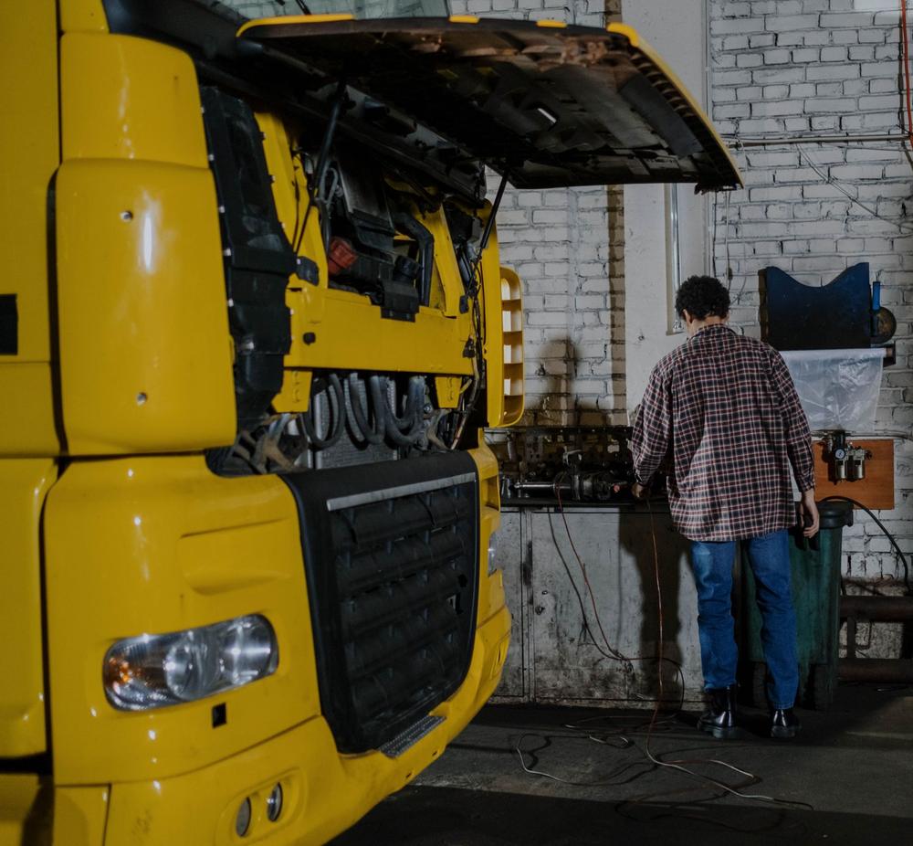 Un camion galben cu cabina ridicată, într-un atelier cu pereți de cărămidă; un om în cămașă în carouri lucrează la bancul de scule.