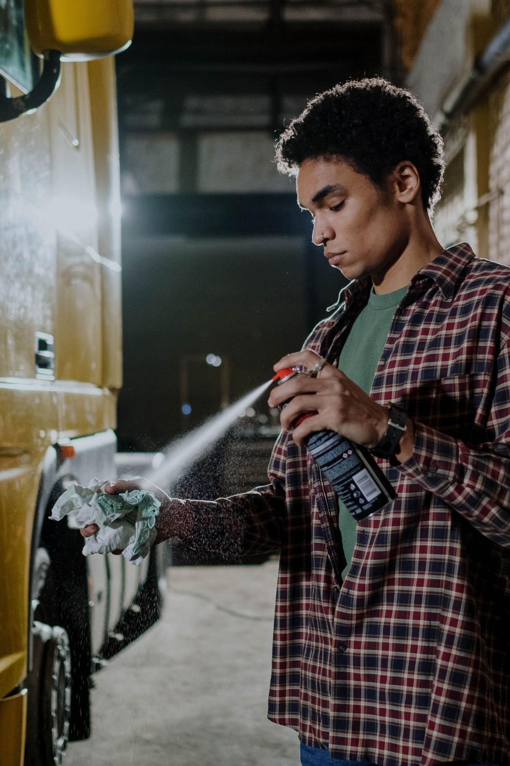 Un om dă cu spray pe o cârpă, lângă botul unui camion galben, într-un atelier cu pereți de cărămidă.