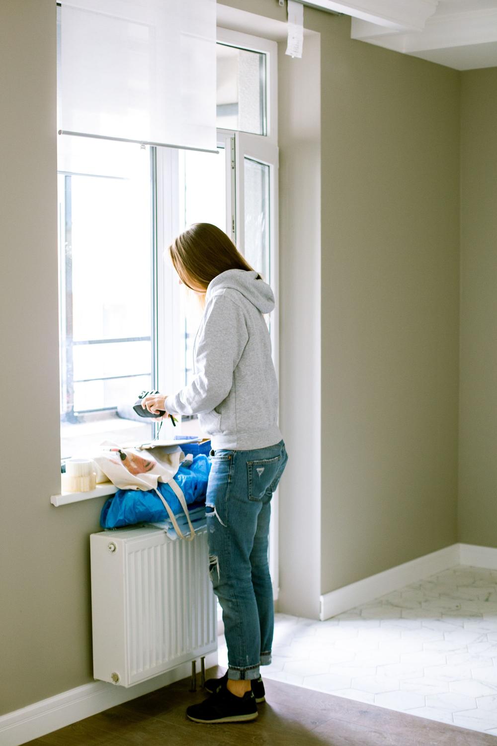 O tânără la fereastra unui apartament gata: pereți zugrăviți kaki, calorifer alb, gresie albă și parchet, o geantă pe pervaz.