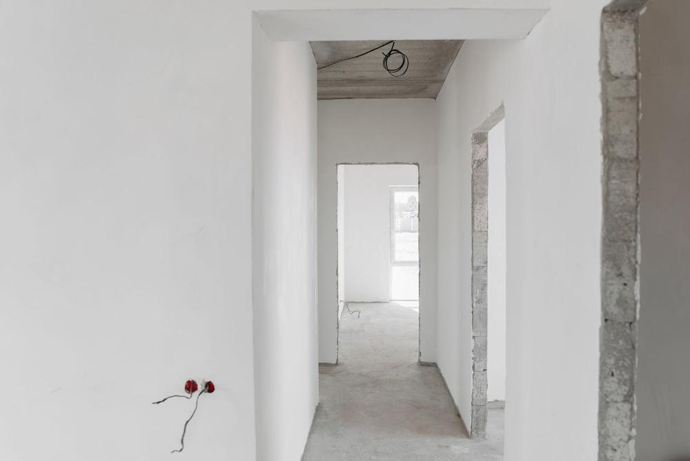 Holul unui apartament la gri: doze roșii jos pe perete, un cablu încolăcit la tavan, goluri de ușă cu marginile de zidărie.