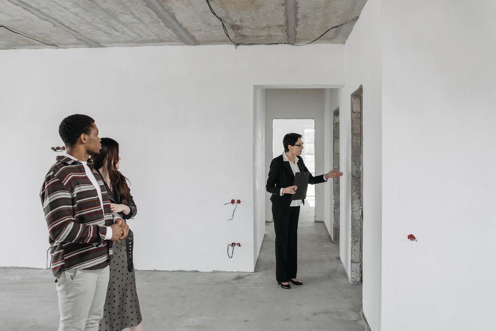 Un cuplu și șefa de șantier, cu un clipboard, într-un apartament la gri: tavan de beton cu cabluri prinse, pereți albi, doze roșii fără prize.