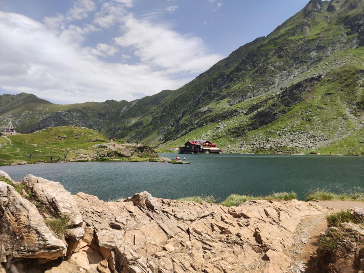 Lacul Bâlea între versanți verzi, cu cabana cu acoperiș roșu pe mal și stânci în prim-plan