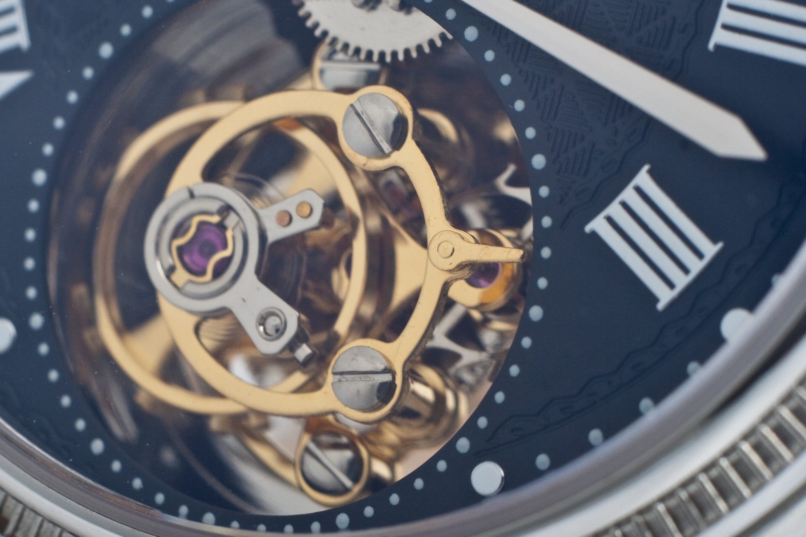 Macro photograph of a gold flying tourbillon cage on a blue watch dial with roman numerals