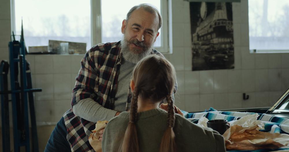 Ilie și fata lui mănâncă pe capota mașinii, pe o pătură cu dungi.