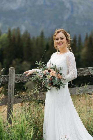 Mireasa cu buchetul de flori sprijinită de un gard de lemn, cu munții în spate