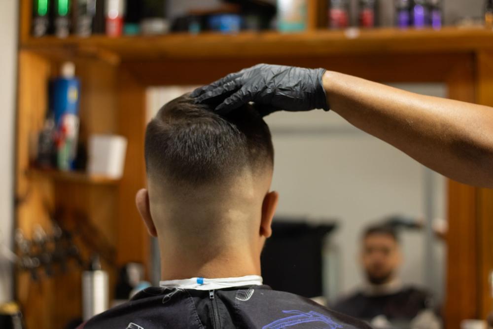 O mână în mănușă neagră verifică părul de sus; clientul se vede în oglindă, în pelerină.