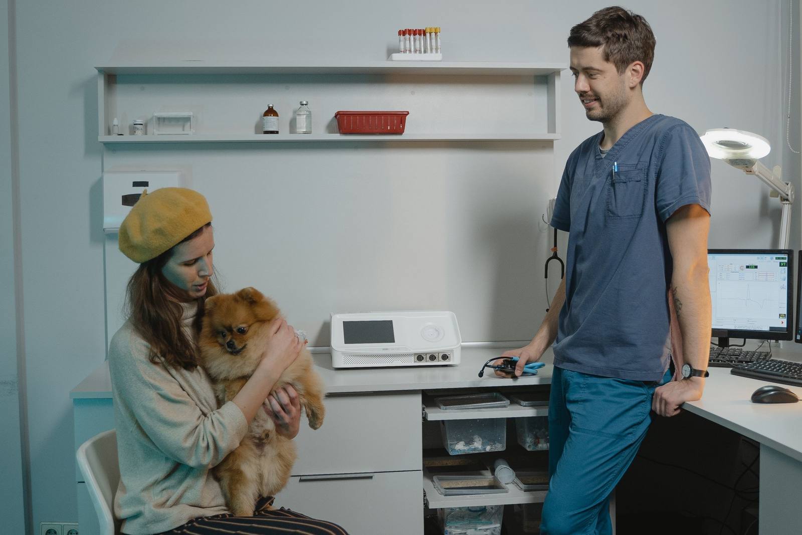 O stăpână cu beretă muștar ține în brațe un pomeranian roșcat, pe scaun; medicul, în uniformă bleumarin, stă lângă ea, sprijinit de birou, și zâmbește.