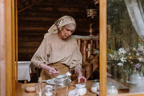 Gazda, cu basma și bluză de in, pune zahăr într-un borcan la o masă de lemn, lângă o fereastră cu flori de câmp într-un bidon