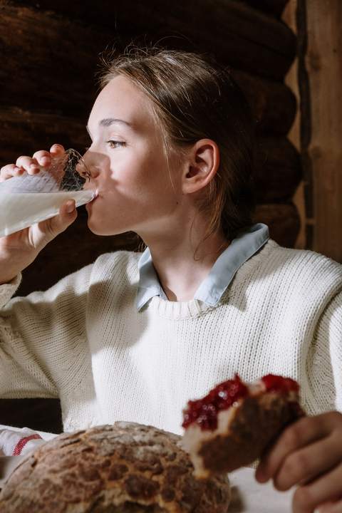 O fată în pulover alb bea lapte dintr-un pahar, cu o felie de pâine cu dulceață în mână