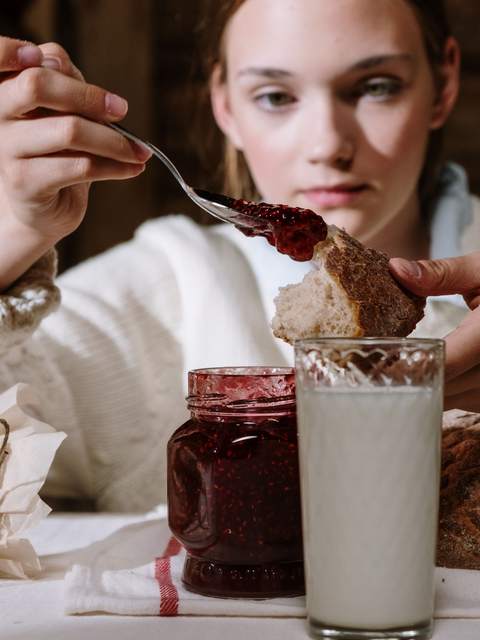 O fată în pulover alb pune dulceață de zmeură pe o felie de pâine; în față, borcanul și un pahar cu lapte