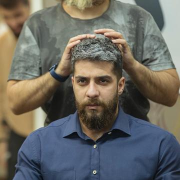 Un bărbat cu barbă neagră și cămașă albastră; frizerul îi ține palmele pe părul grizonat.