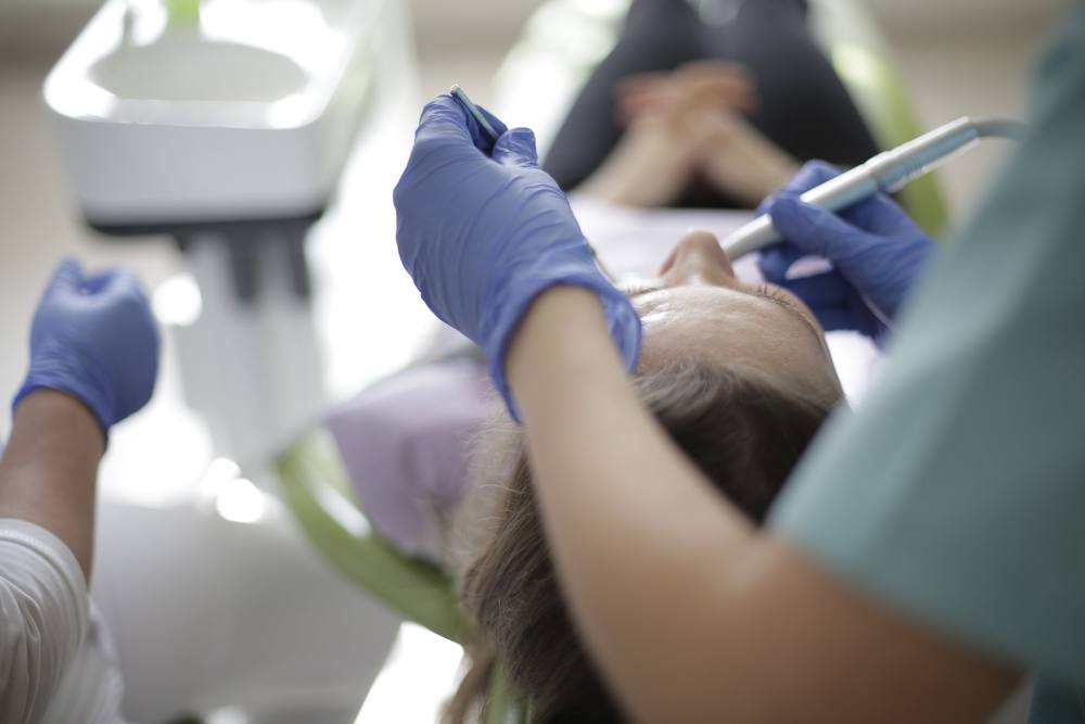 Prim-plan cu mâinile medicului în mănuși albastre, cu oglinda și turbina, deasupra unei paciente întinse.