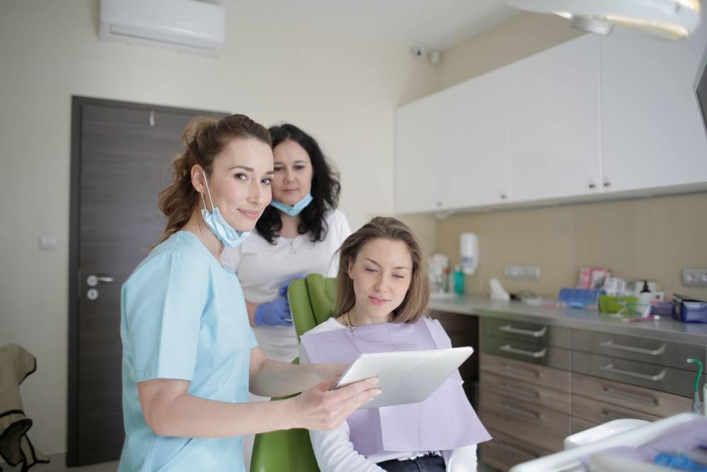 O medic arată pe o tabletă planul de tratament unei paciente care stă pe scaunul verde; colega ei se uită peste umăr.