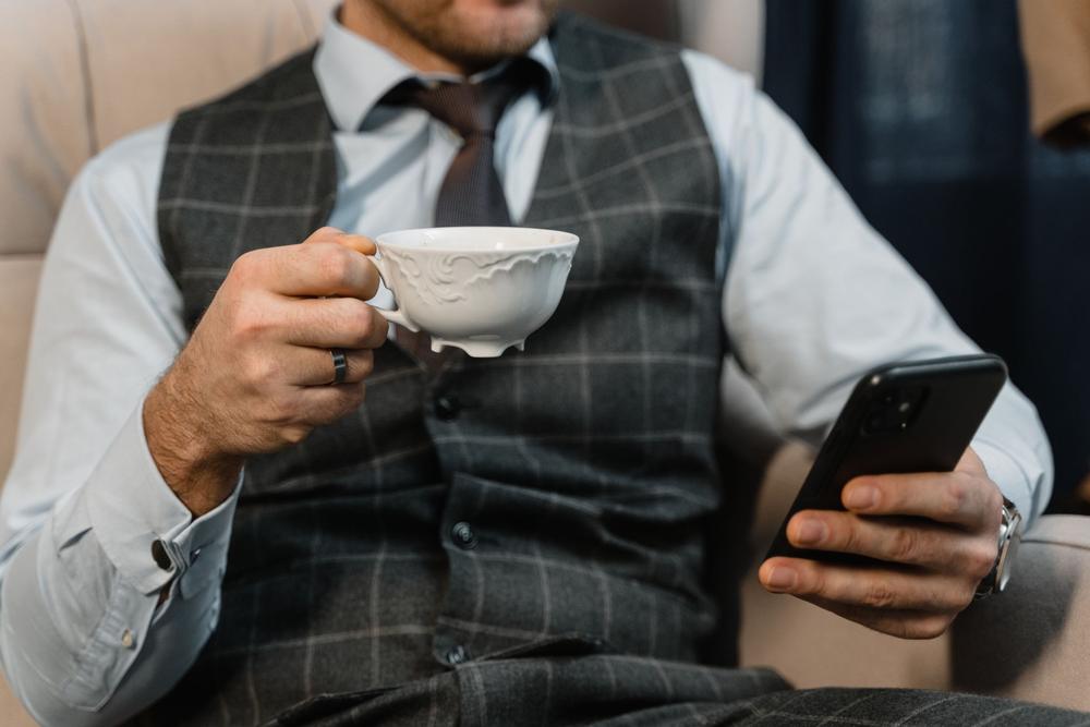 Un client în vestă în carouri ține o ceașcă albă de porțelan într-o mână și telefonul în cealaltă, așezat pe o canapea bej.