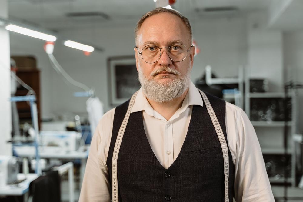 Un croitor cu barbă albă și ochelari rotunzi, în cămașă albă și vestă închisă, cu centimetrul pe după gât, privește drept în aparat; în spate, atelierul cu neoane.