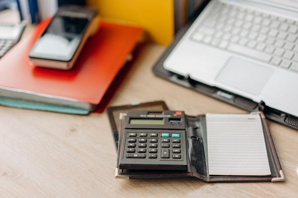 Calculator de birou pe o agendă cu foi liniate; în spate, dosare roșii și galbene și un laptop pe suport.